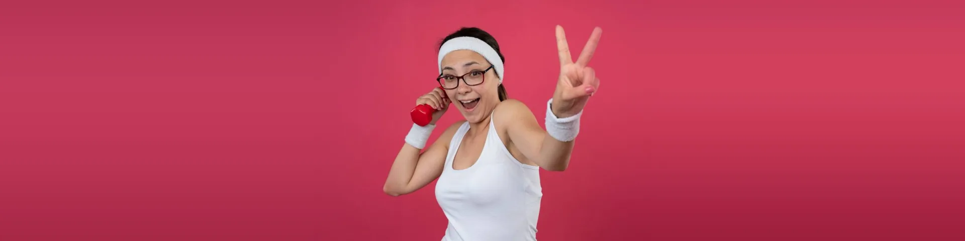Junge Frau in Sportkleidung vor pinkem Hintergrund freut sich mit Victory-Zeichen und Hantel