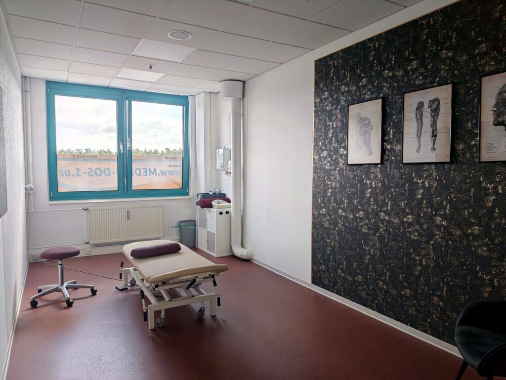 Behandlungsraum mit Liege, Fenster und anatomischen Bildern